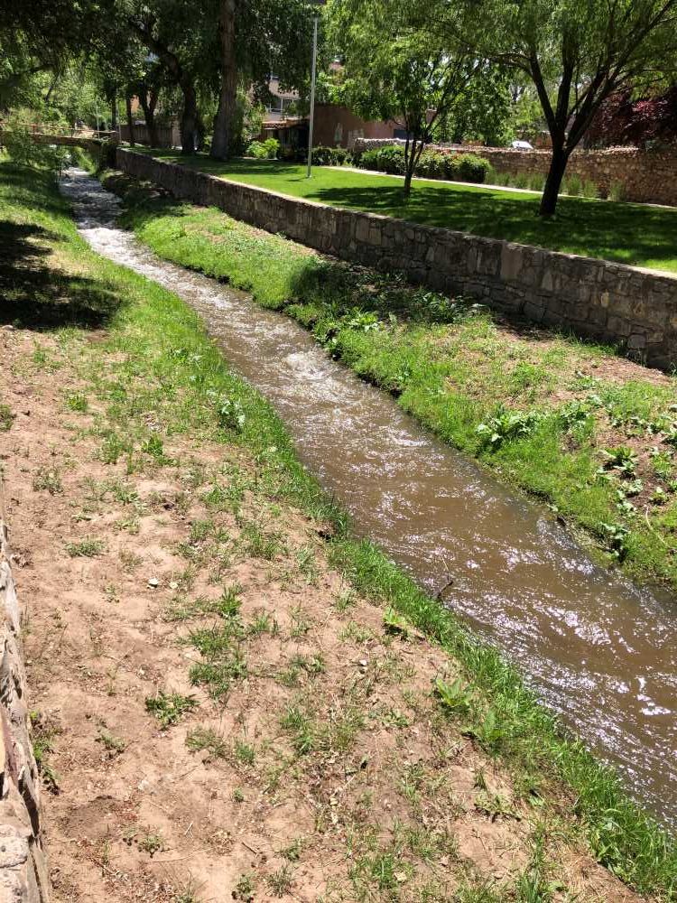 The Santa Fe River meanders through a park, bordered by a grassy bank. Sunlight filters through trees, casting shadows on the water.