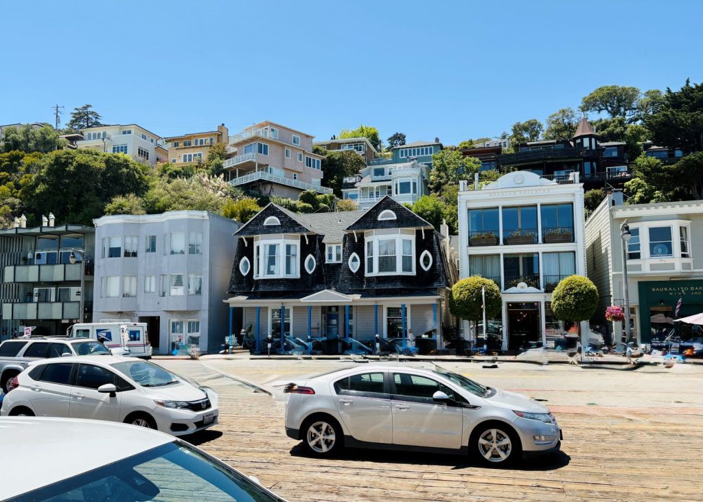 Charming coastal town street with colorful buildings and parked cars. Blue sky, sunny day, and lush hillside create a lively, inviting atmosphere.