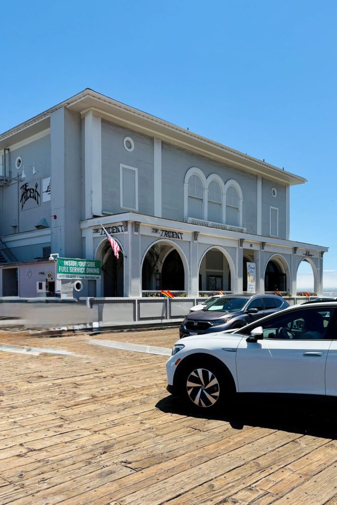 A large building with arched windows, labeled "The Trident," sits on a wooden pier under a clear blue sky. Cars are parked in front of this seaside restaurant.