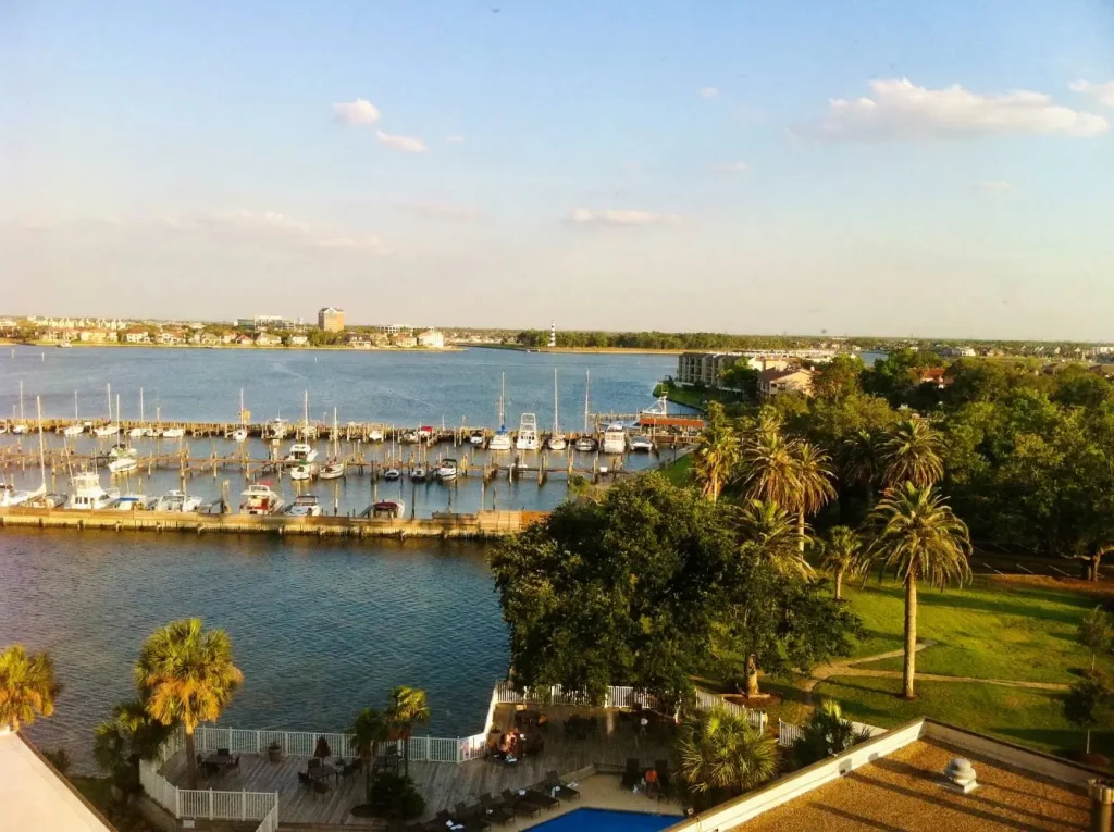 A serene harbor view of Clear Lake, TX with docked boats, lush trees and a clear sky. The scene conveys a peaceful, idyllic coastal setting.