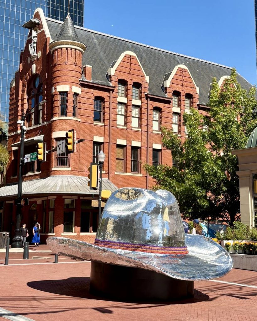 A large bejeweled cowboy hat sculpture sits in Fort Worth's Sundance Square against a historic red brick building with arched windows. The scene is bright and sunny.