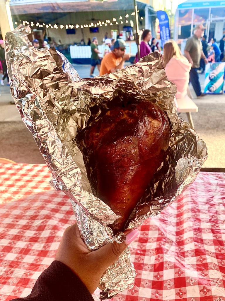 A hand holds a roasted turkey leg wrapped in foil at the State Fair of Texas. The red-and-white checkered tablecloth and bustling background suggest a lively, casual atmosphere.