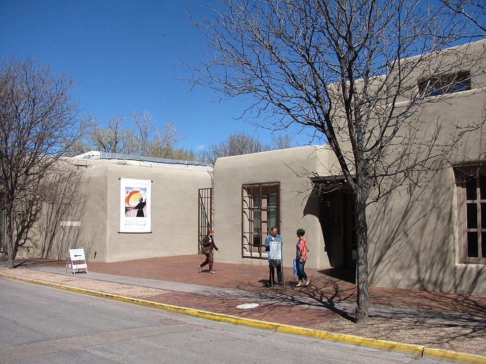 The Georgia O'Keefe Museum in an adobe-style building with people walking and tree shadows on the walls. A colorful art poster hangs outside, creating a calm, sunny scene.