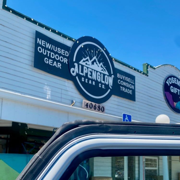Storefront for "Alpenglow Gear Co." with bold signage offering new/used outdoor gear and trade options. A car's window frame is visible in the foreground.