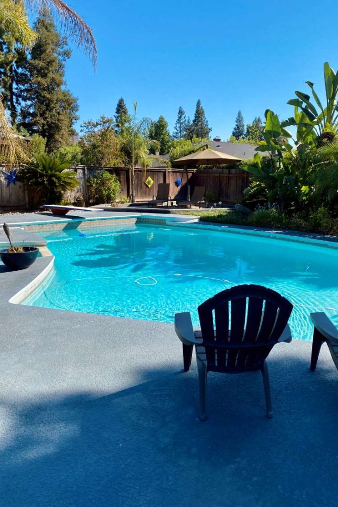 A tranquil backyard scene at an AirBnB in for a day trip to Yosemite. A clear blue swimming pool and two empty Adirondack chairs surrounded by greenery and a wooden fence.
