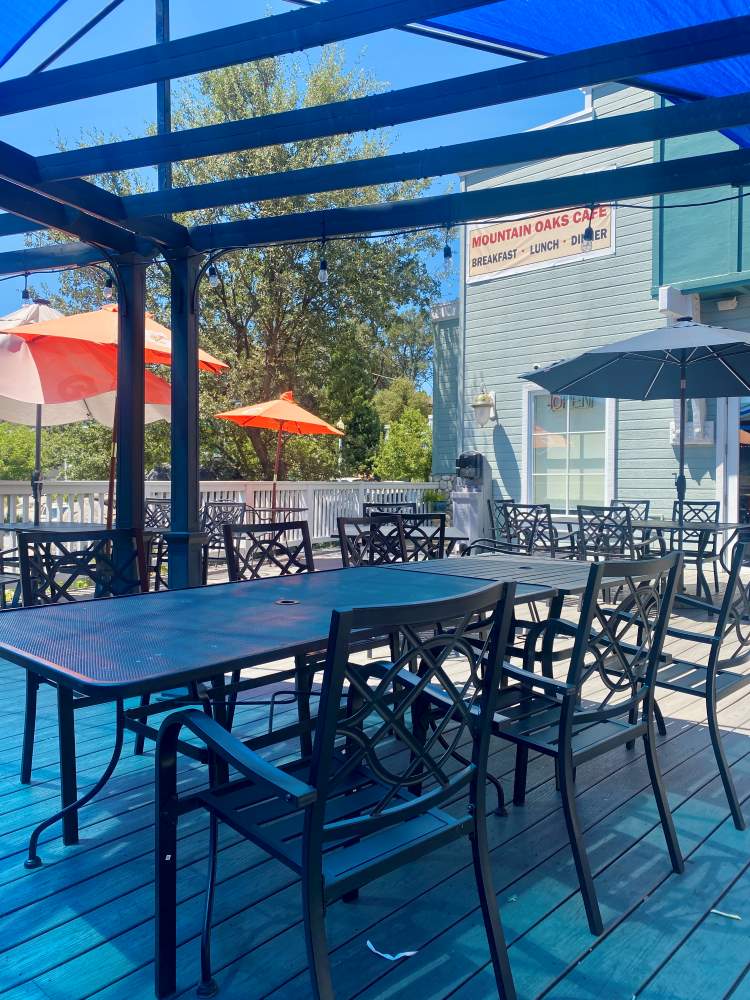 Outdoor seating area of Mountain Oaks Café with dark metal tables and chairs under a blue pergola. Bright orange umbrellas and trees add a vibrant touch.