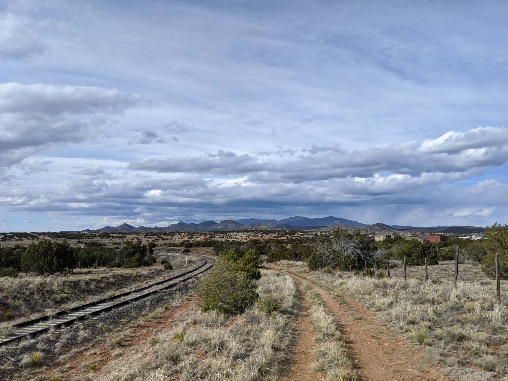 A scenic view of the Sangre de Cristo mountains with a dirt path and railway track running parallel, leading to distant mountains under a cloudy sky. The scene feels serene and open.