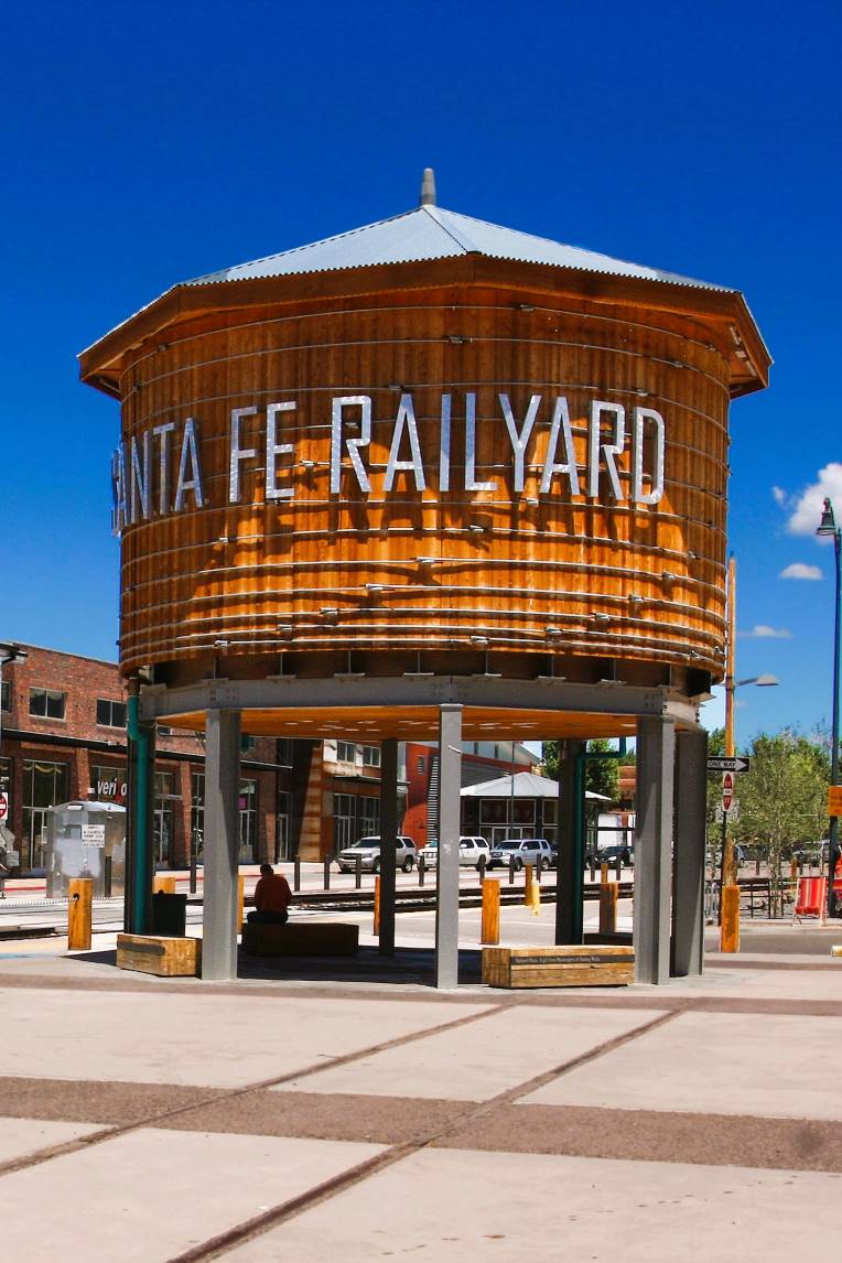 Large wooden water tower at Santa Fe Railyard on a sunny day. People sit beneath it, surrounded by a mix of industrial and modern architecture.
