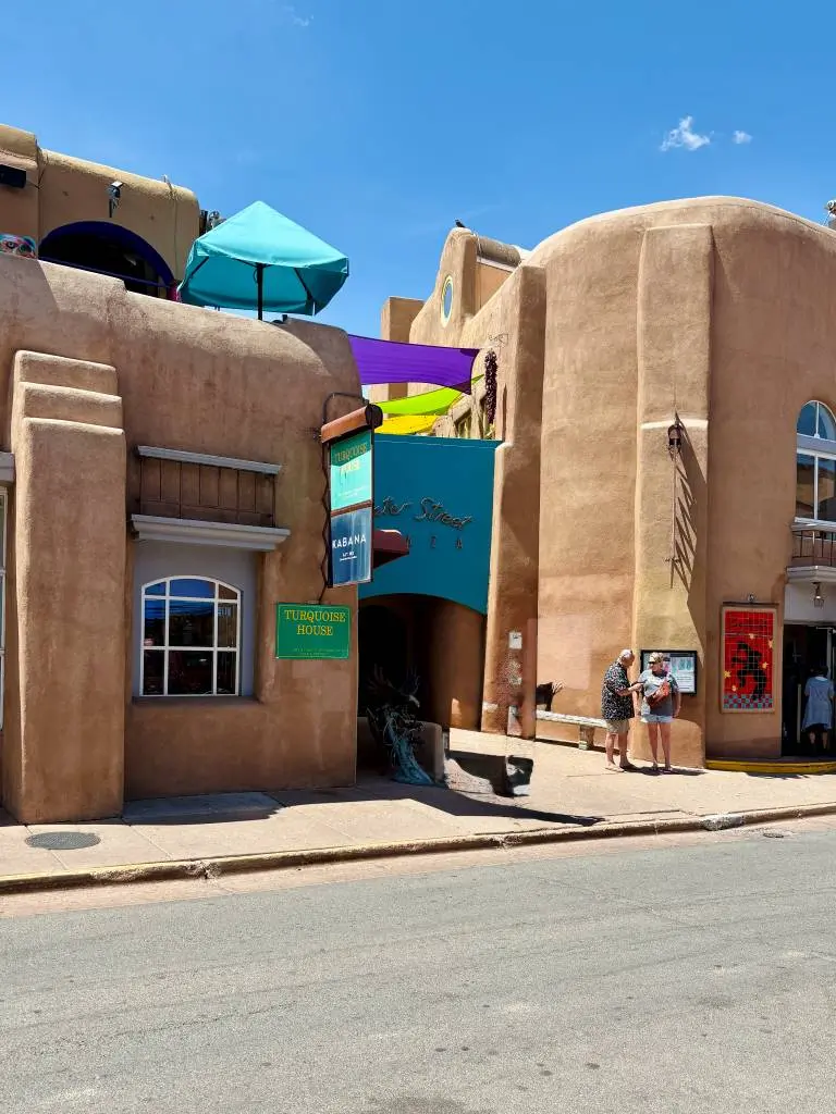 Water Street Plaza's adobe-style building in Santa Fe with bright sunlight, colorful umbrellas and signs. Two shoppers stand talking.