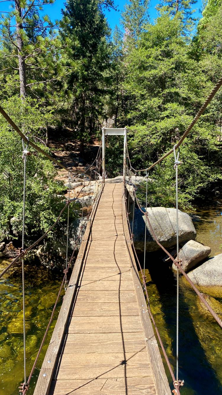 Wooden suspension bridge at Yosemite Park over a clear, rocky stream, surrounded by lush green trees. Sunlight filters through, creating a serene, inviting scene.